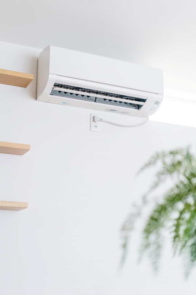 Climatiseur mural blanc design pour intérieur moderne Climatiseur mural blanc installé sur un mur épuré avec étagères en bois et une plante verte en avant-plan.