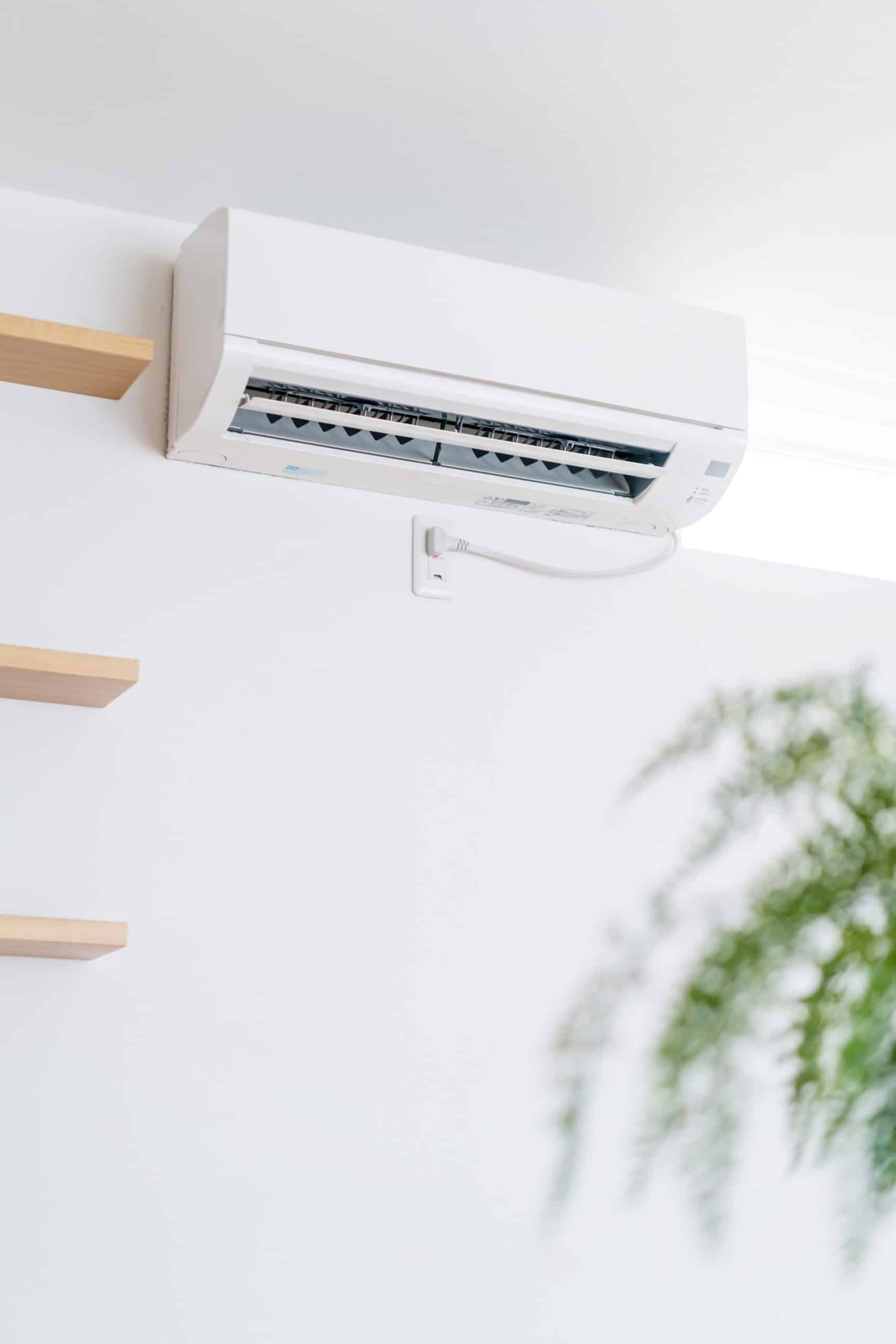 Climatiseur mural blanc installé sur un mur épuré avec étagères en bois et une plante verte en avant-plan.